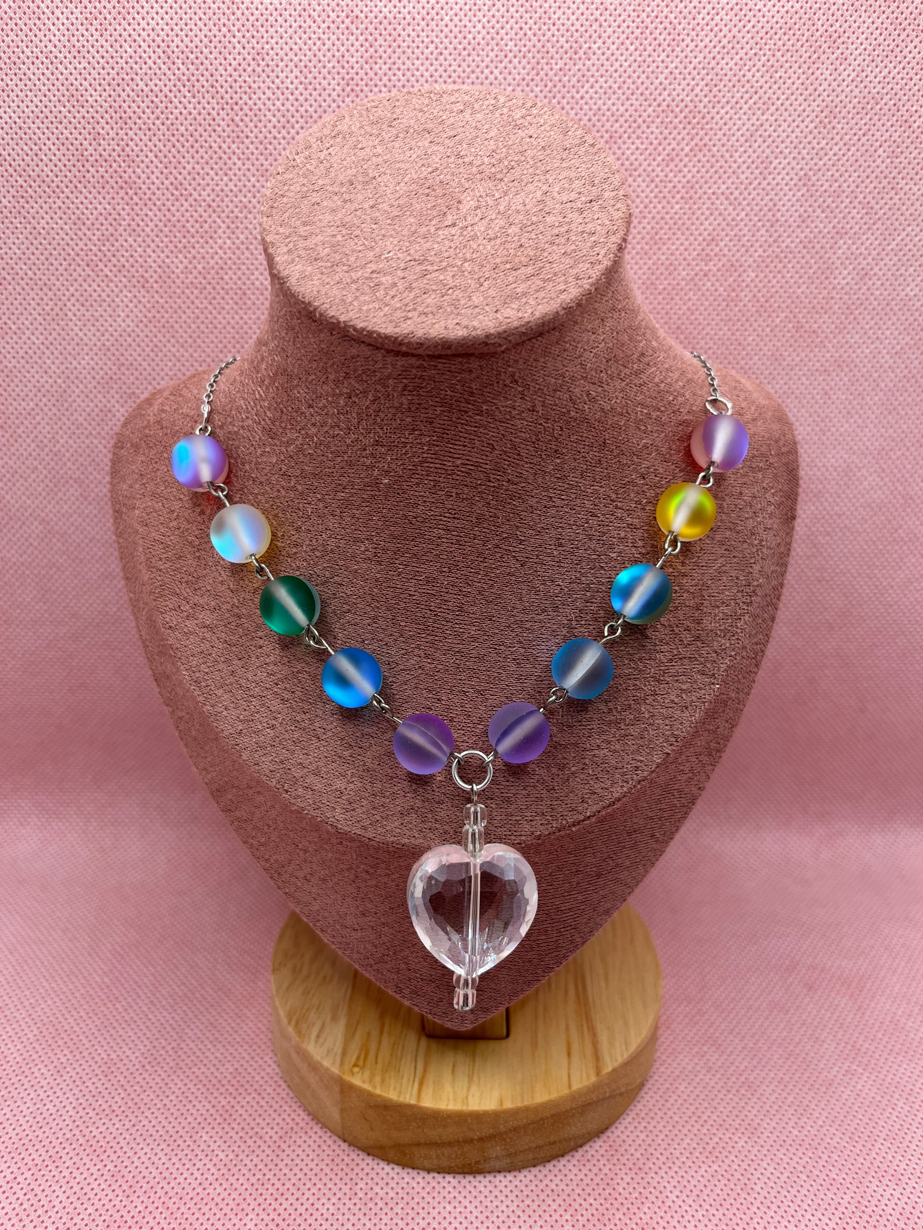 Rainbow reflective beaded necklace with a clear heart-shaped pendant draped over a blush pink mannequin with a pink background.