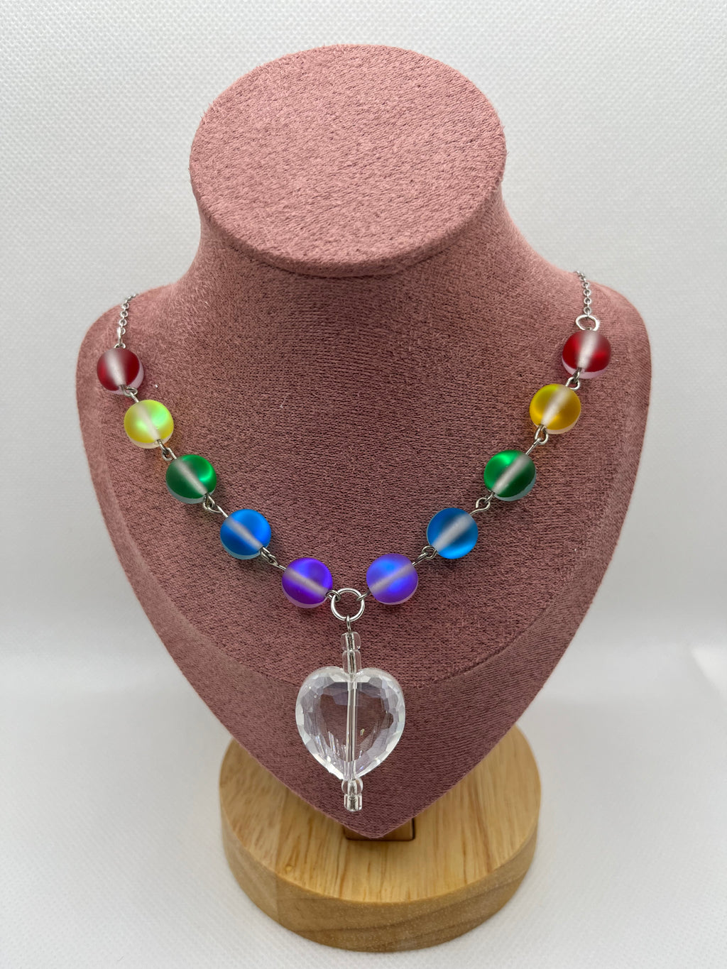 Rainbow reflective beaded necklace with a clear heart-shaped pendant draped over a blush pink mannequin with a white background.