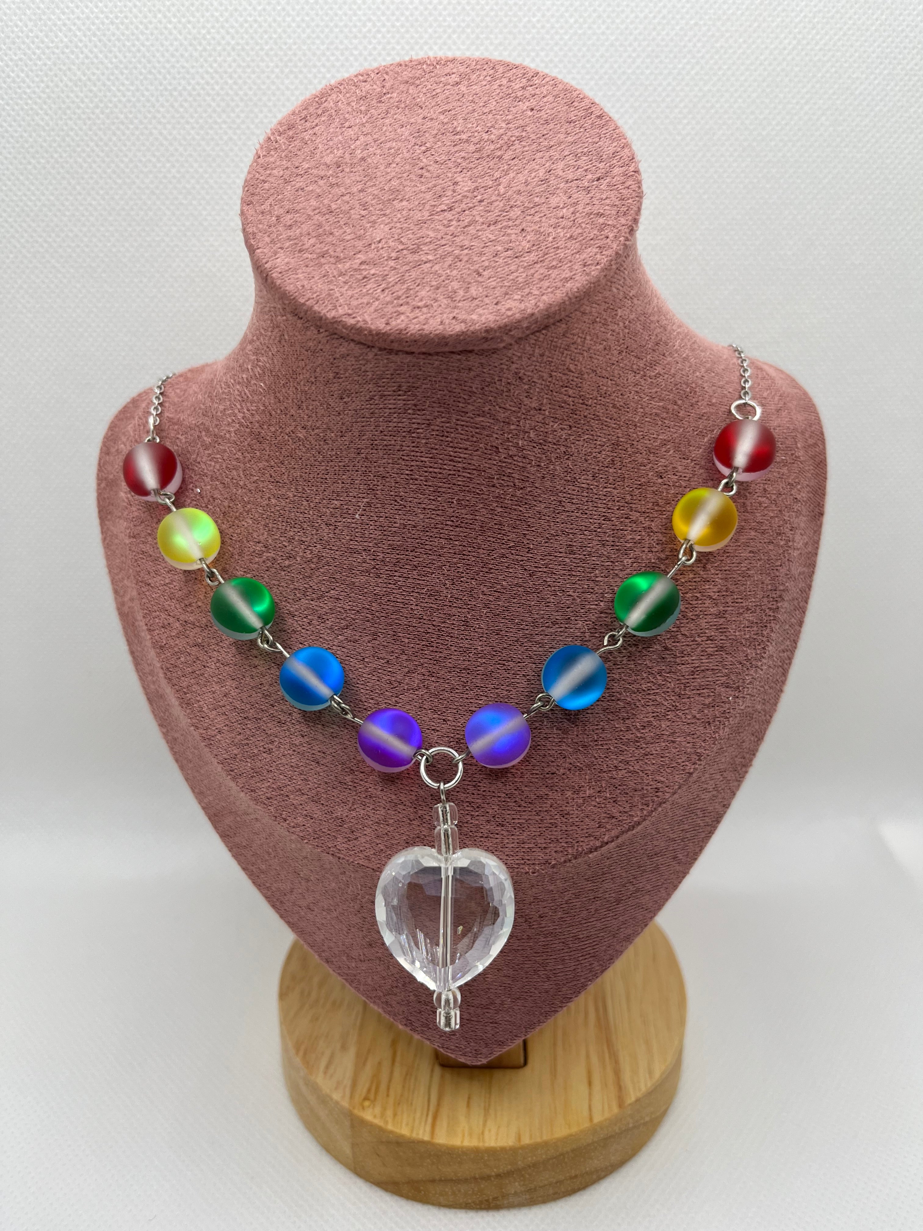 Rainbow reflective beaded necklace with a clear heart-shaped pendant draped over a blush pink mannequin with a white background.