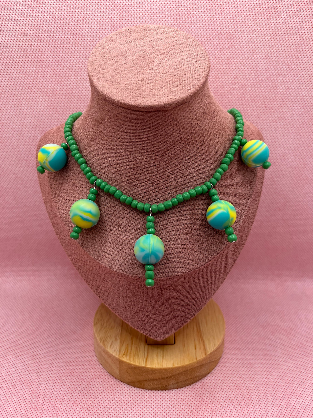 Green seed bead necklace with silicone beads strung throughout draped over a blush pink mannequin with a pink background.