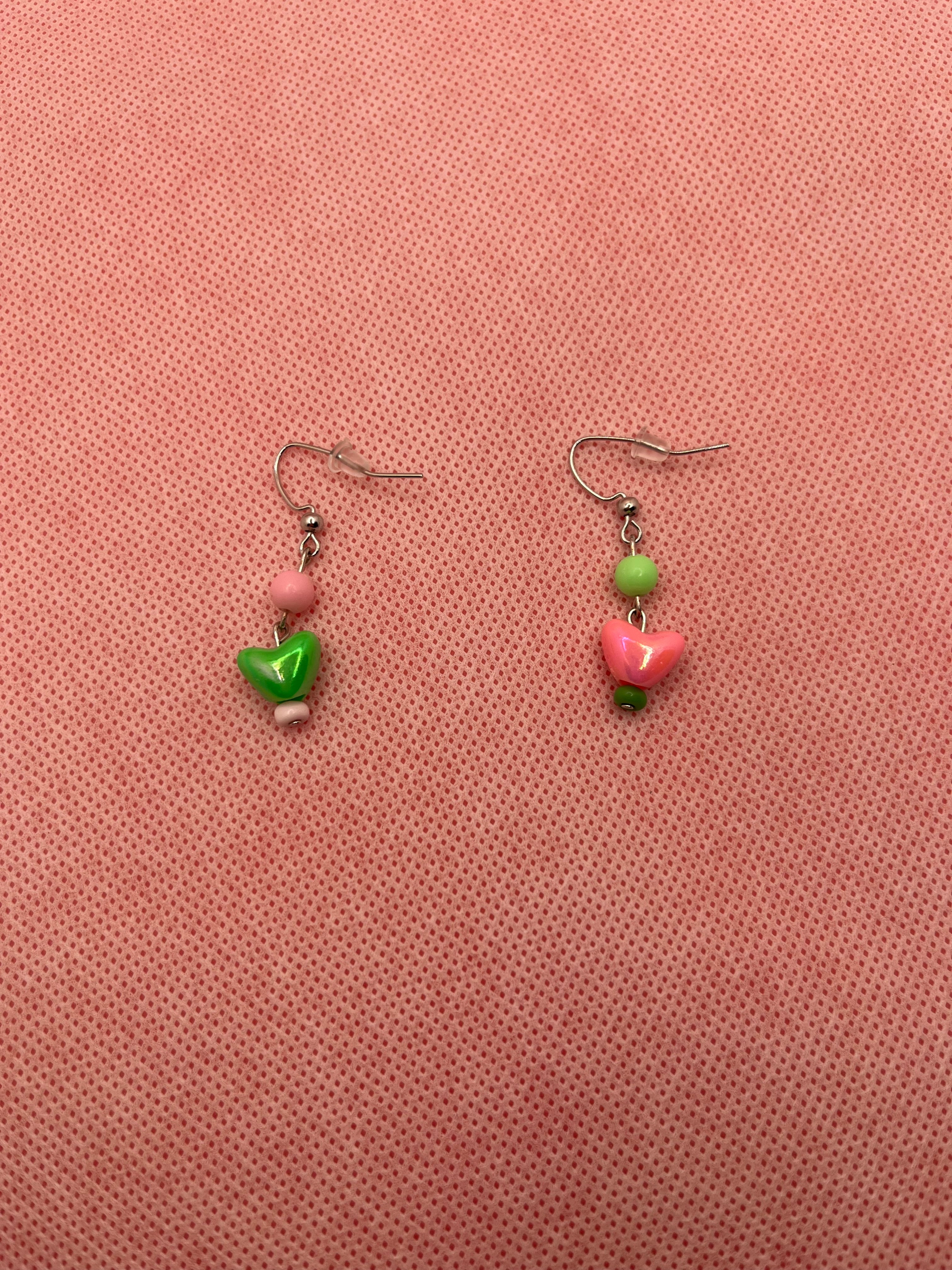 Pink and green alternate earrings with hearts on the end against a pink background