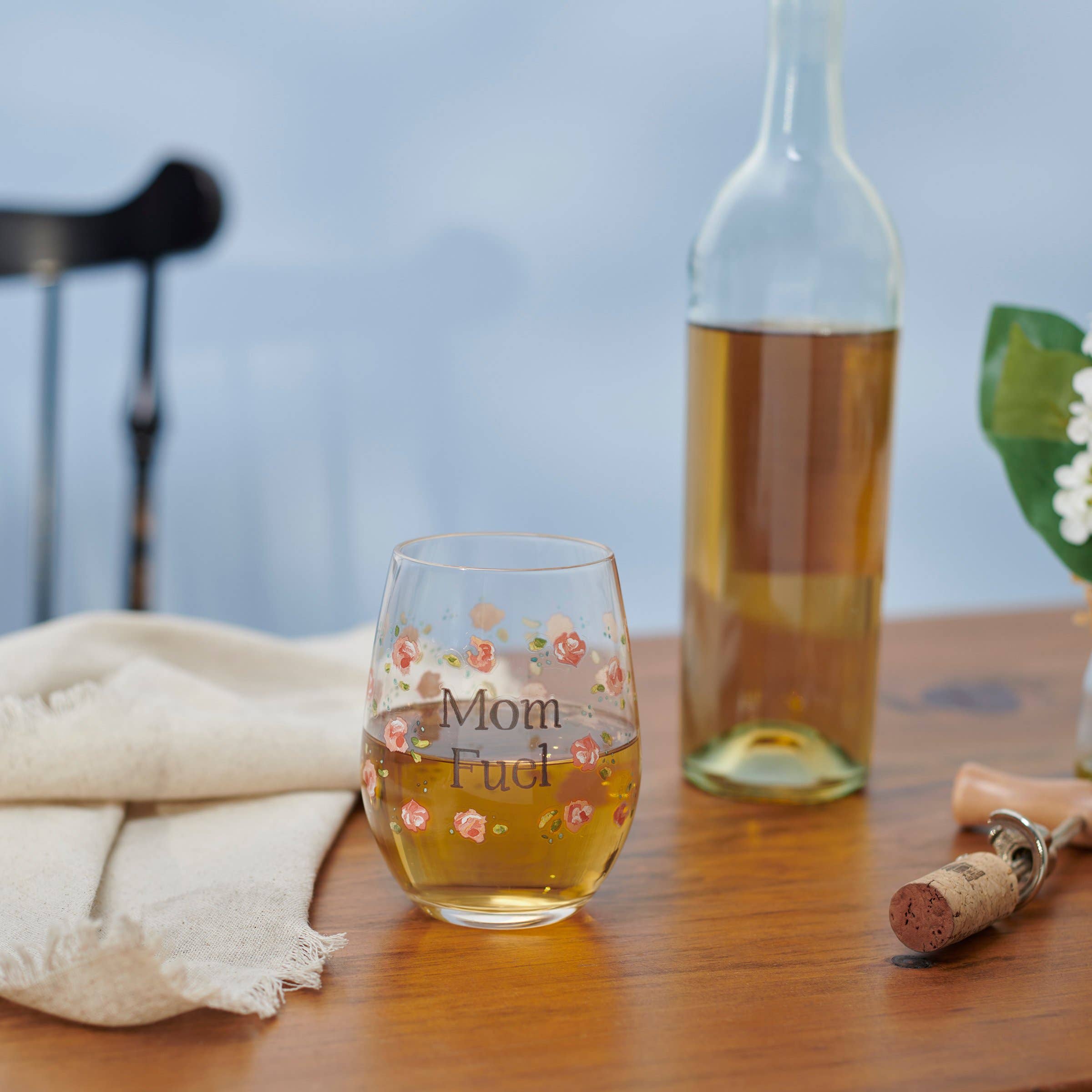 Stemless wine glass with 'Mom Fuel' text on a wooden table with a bottle of wine in the background.