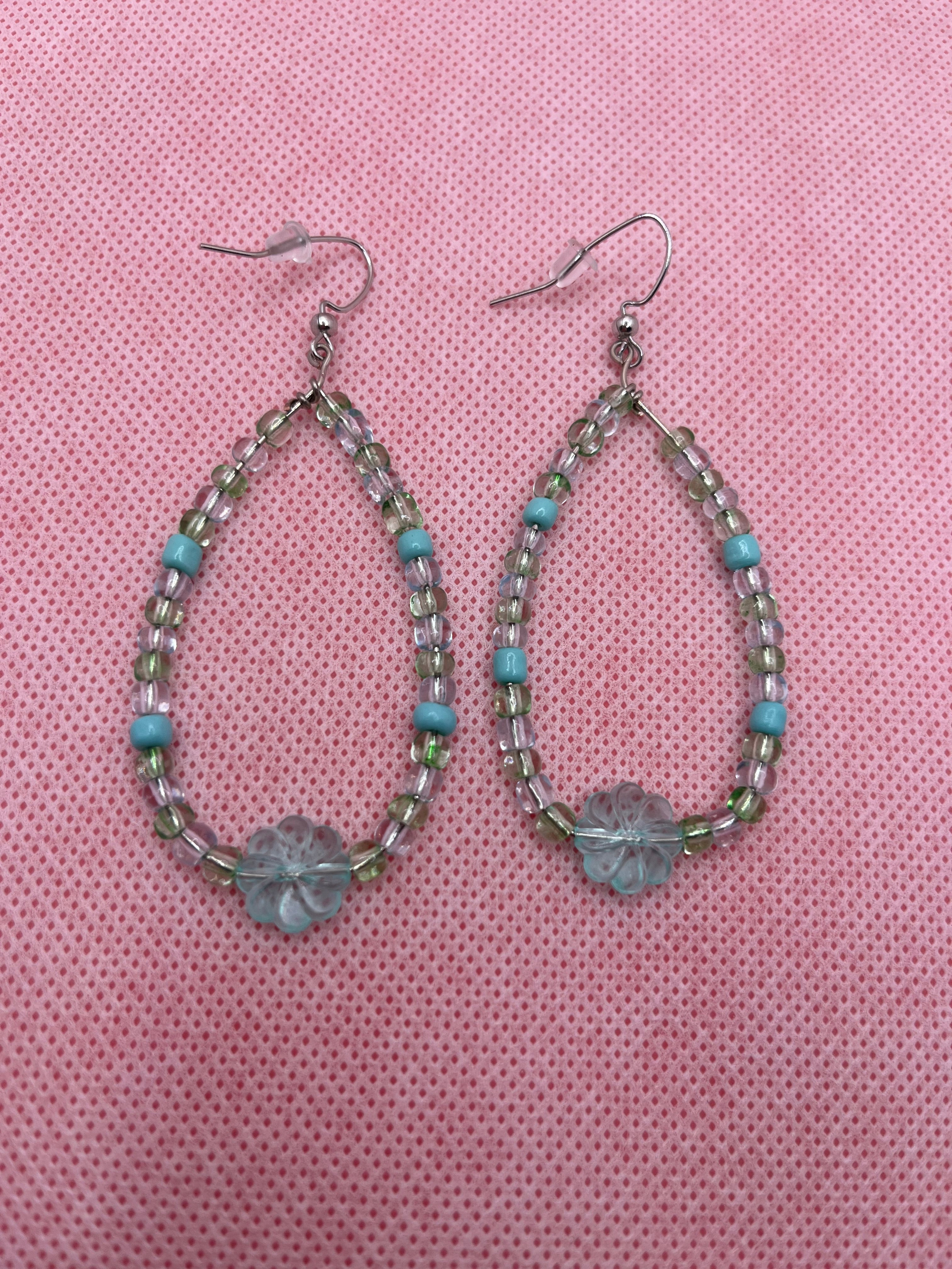 Blue and green beaded earrings with a large transparent clover bead on the bottom against a pink background
