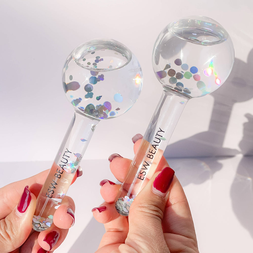 Two clear beauty tools with glitter inside, held by a person with red nail polish on a light background.