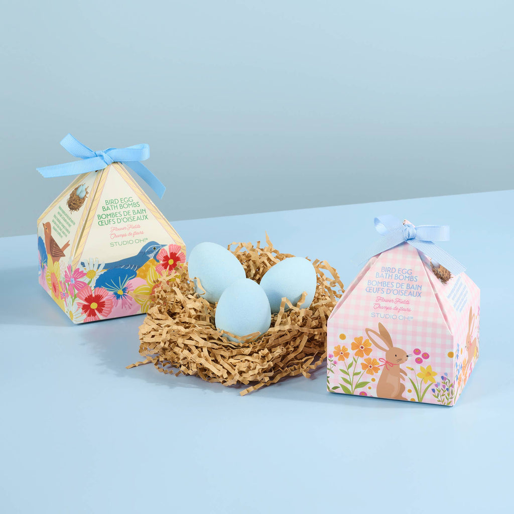 Two egg-themed bath bombs in a nest with their packaging on a light blue background
