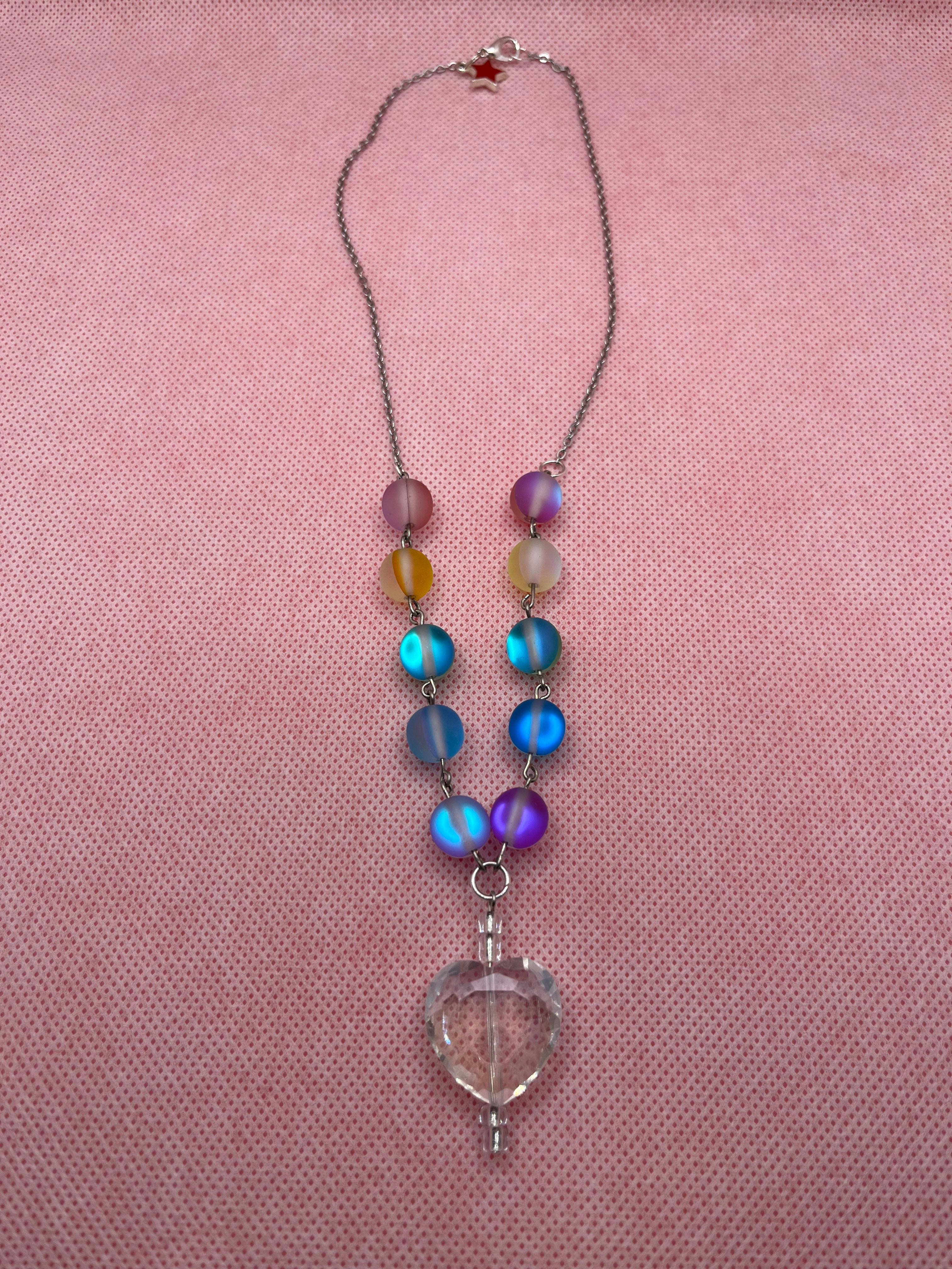 Rainbow reflective beaded necklace with a clear heart-shaped pendant laid flat against a pink background.