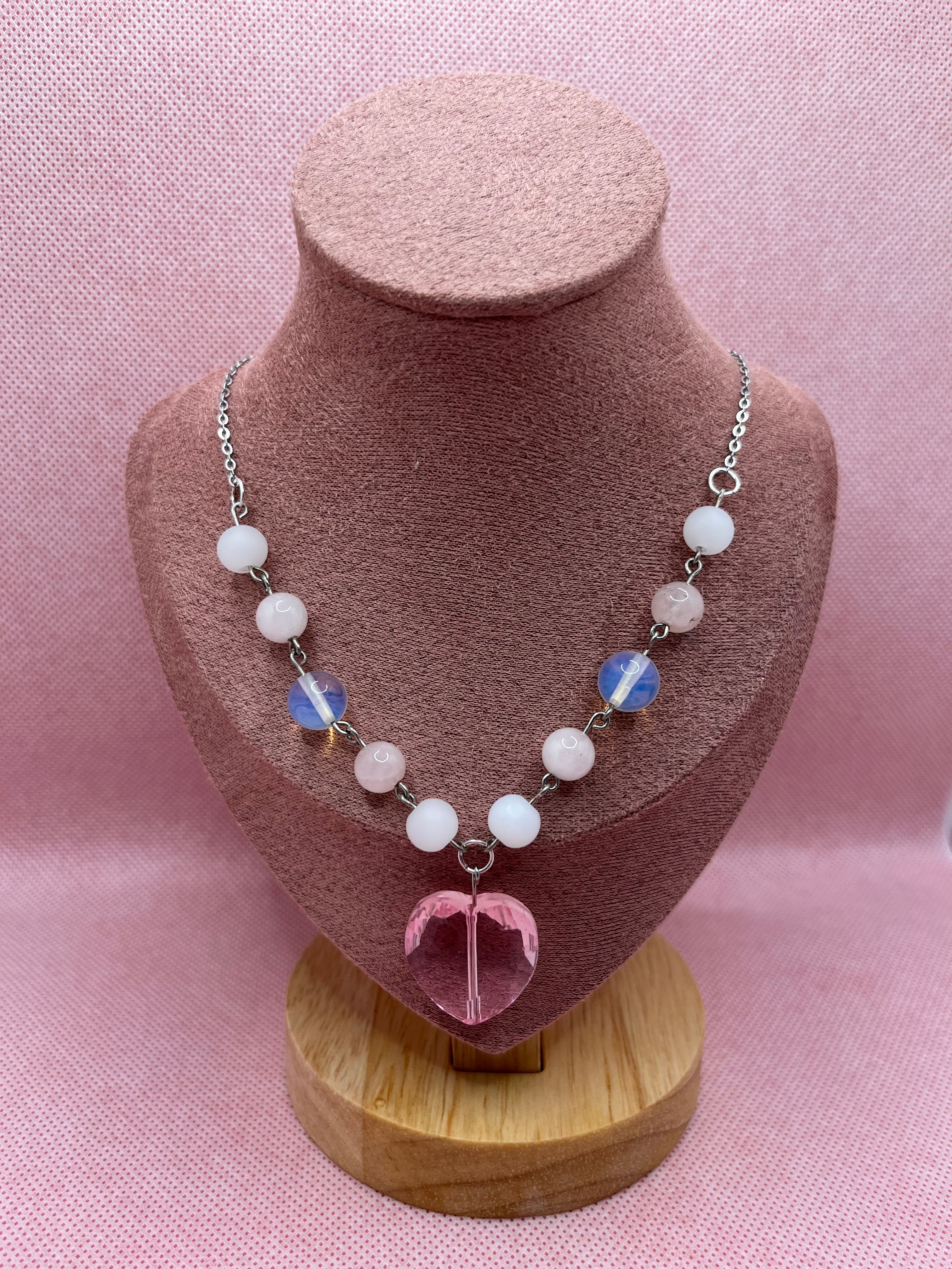 Pink and white necklace with a heart-shaped pendant draped over a blush pink mannequin with a pink background.