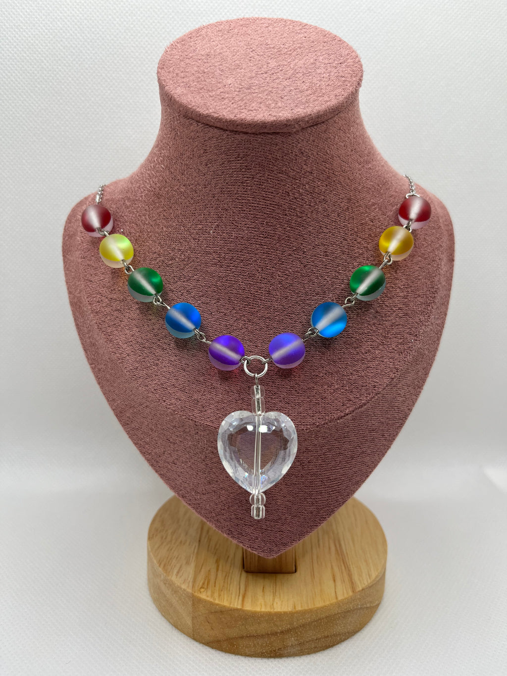 Rainbow reflective beaded necklace with a clear heart-shaped pendant draped over a blush pink mannequin with a white background.