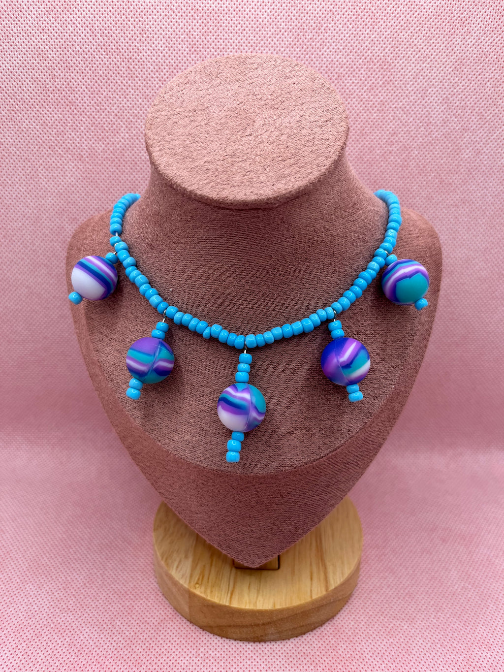 Blue seed bead necklace with silicone beads strung throughout draped over a blush pink mannequin with a pink background.