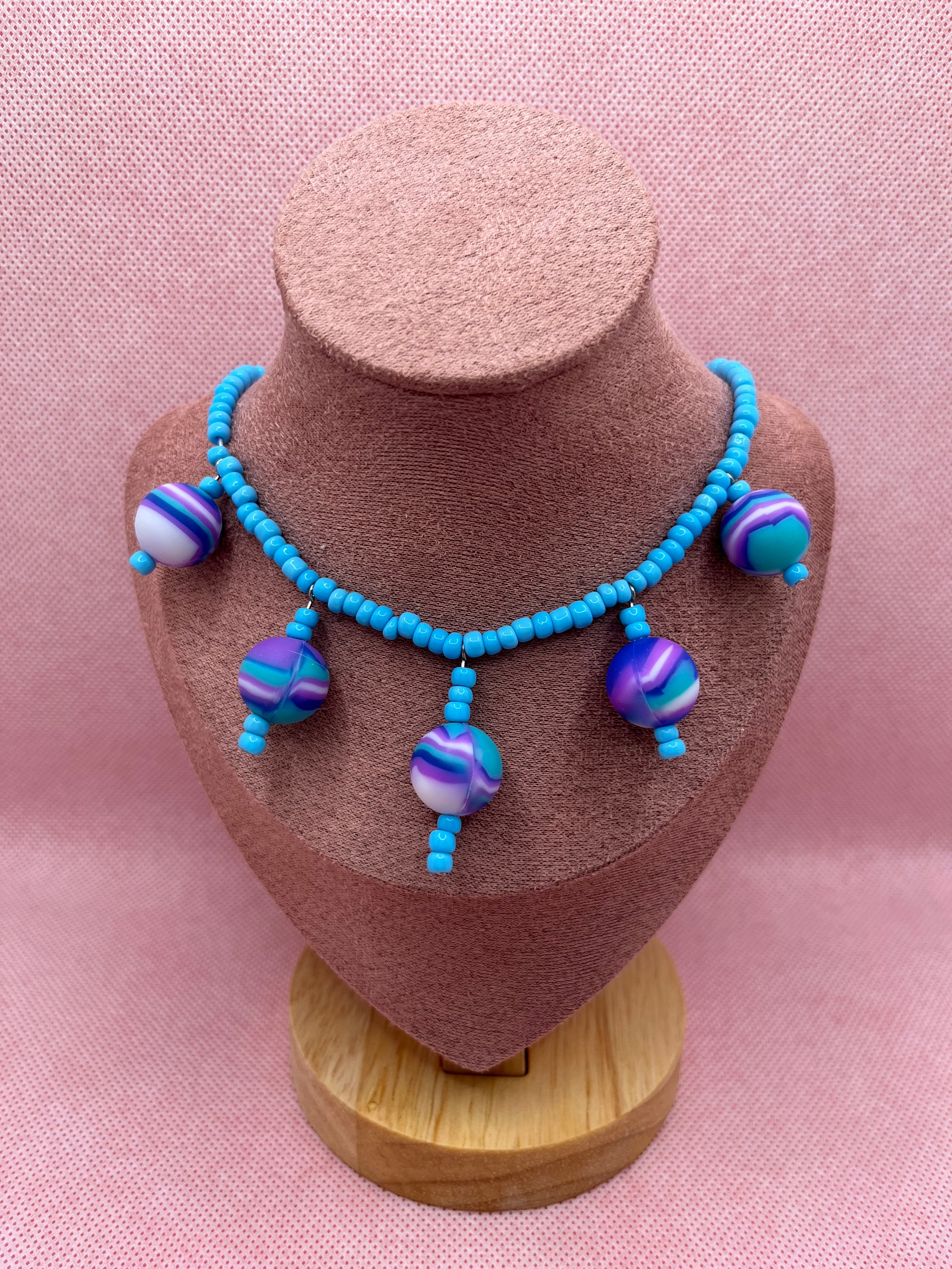 Blue seed bead necklace with silicone beads strung throughout draped over a blush pink mannequin with a pink background.