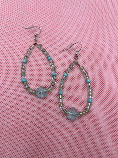 Blue and green beaded earrings with a large transparent clover bead on the bottom against a pink background