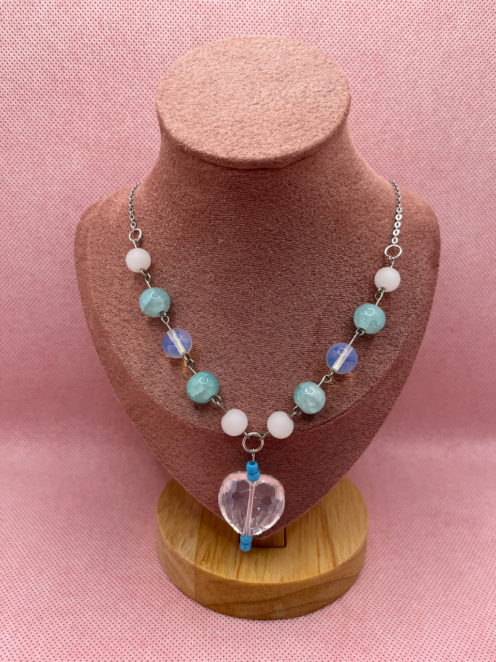 Blue, white, and clear necklace with a heart-shaped pendant draped over a blush pink mannequin with a pink background.