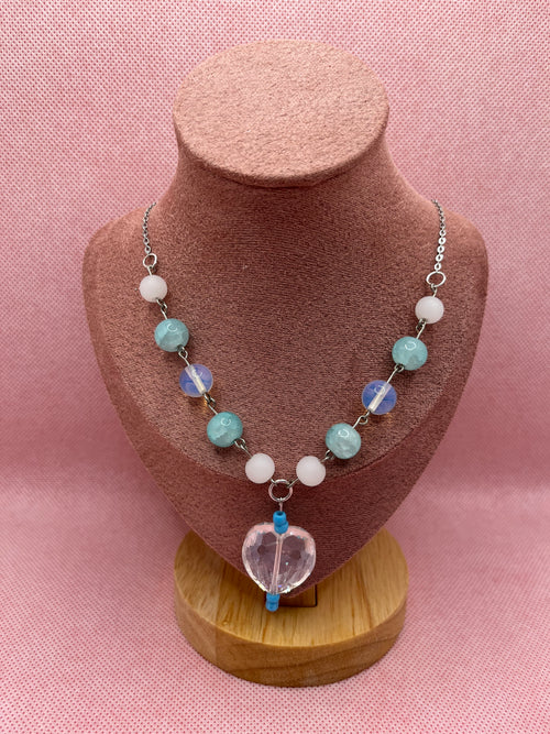 Blue, white, and clear necklace with a heart-shaped pendant draped over a blush pink mannequin with a pink background.