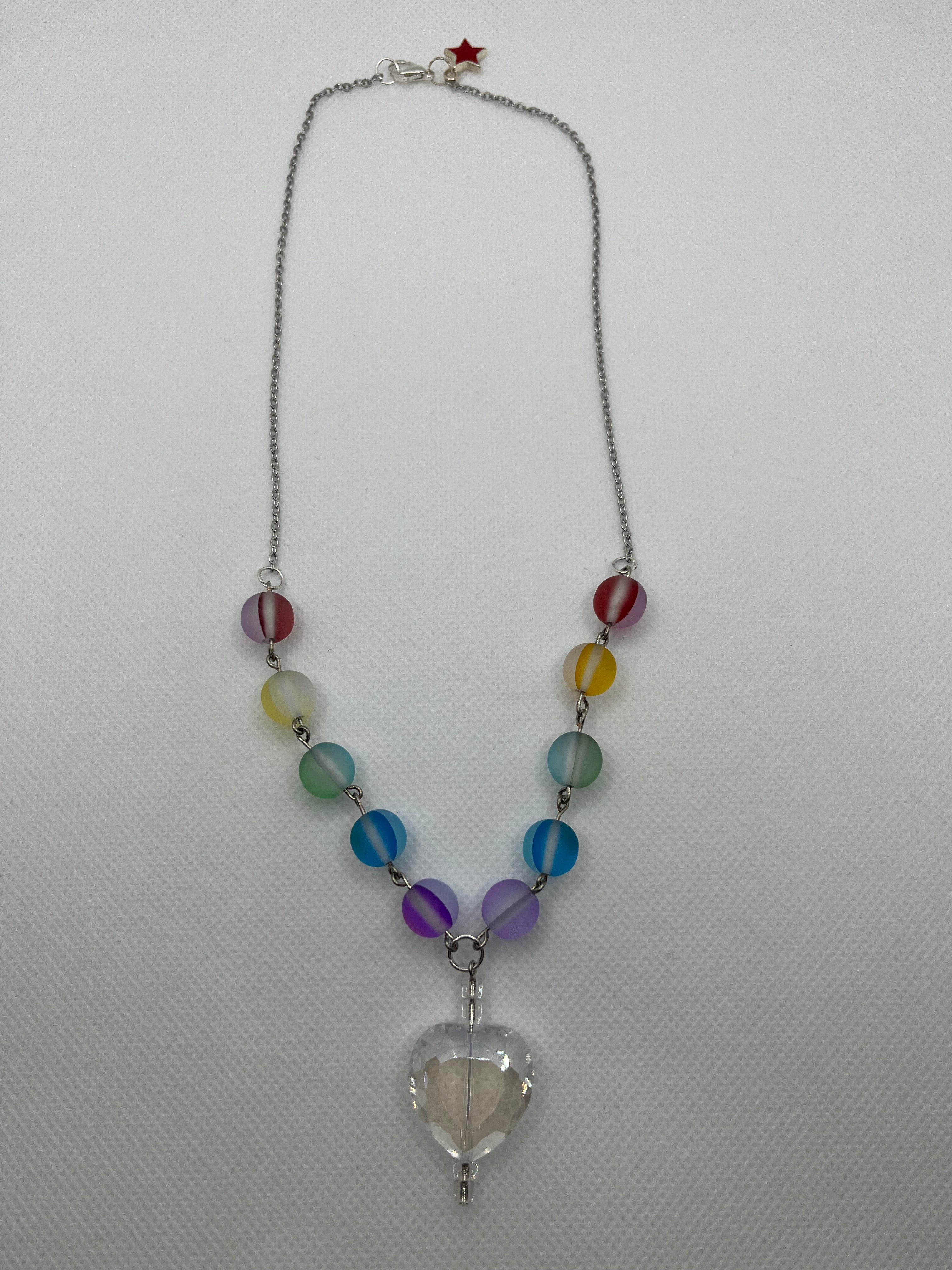 Rainbow reflective beaded necklace with a clear heart-shaped pendant laid flat against a white background.
