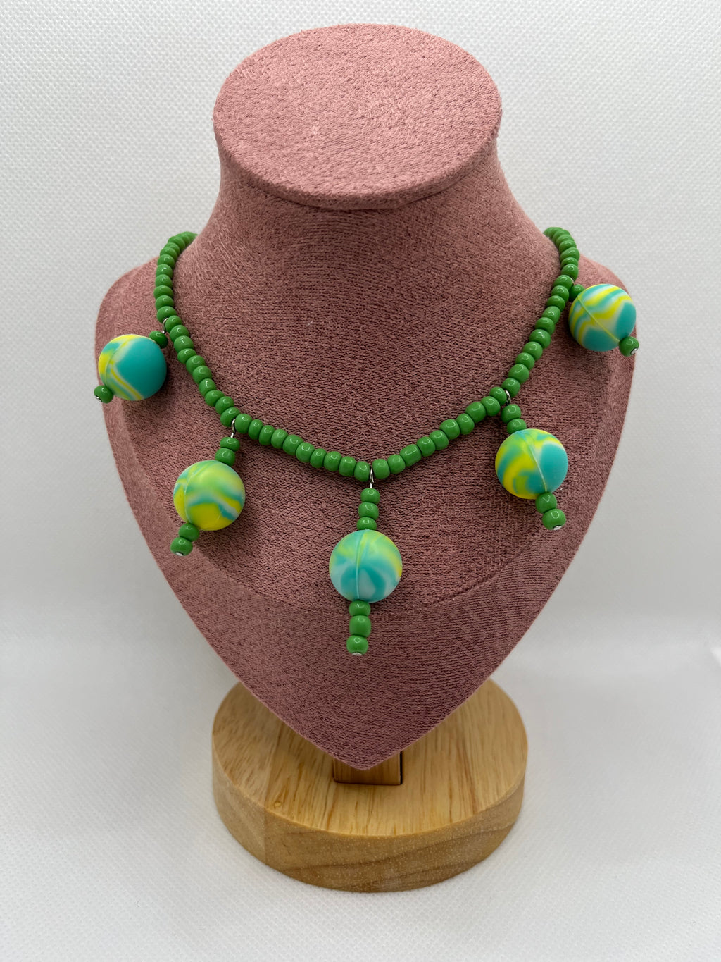 Green seed bead necklace with silicone beads strung throughout draped over a blush pink mannequin with a white background.
