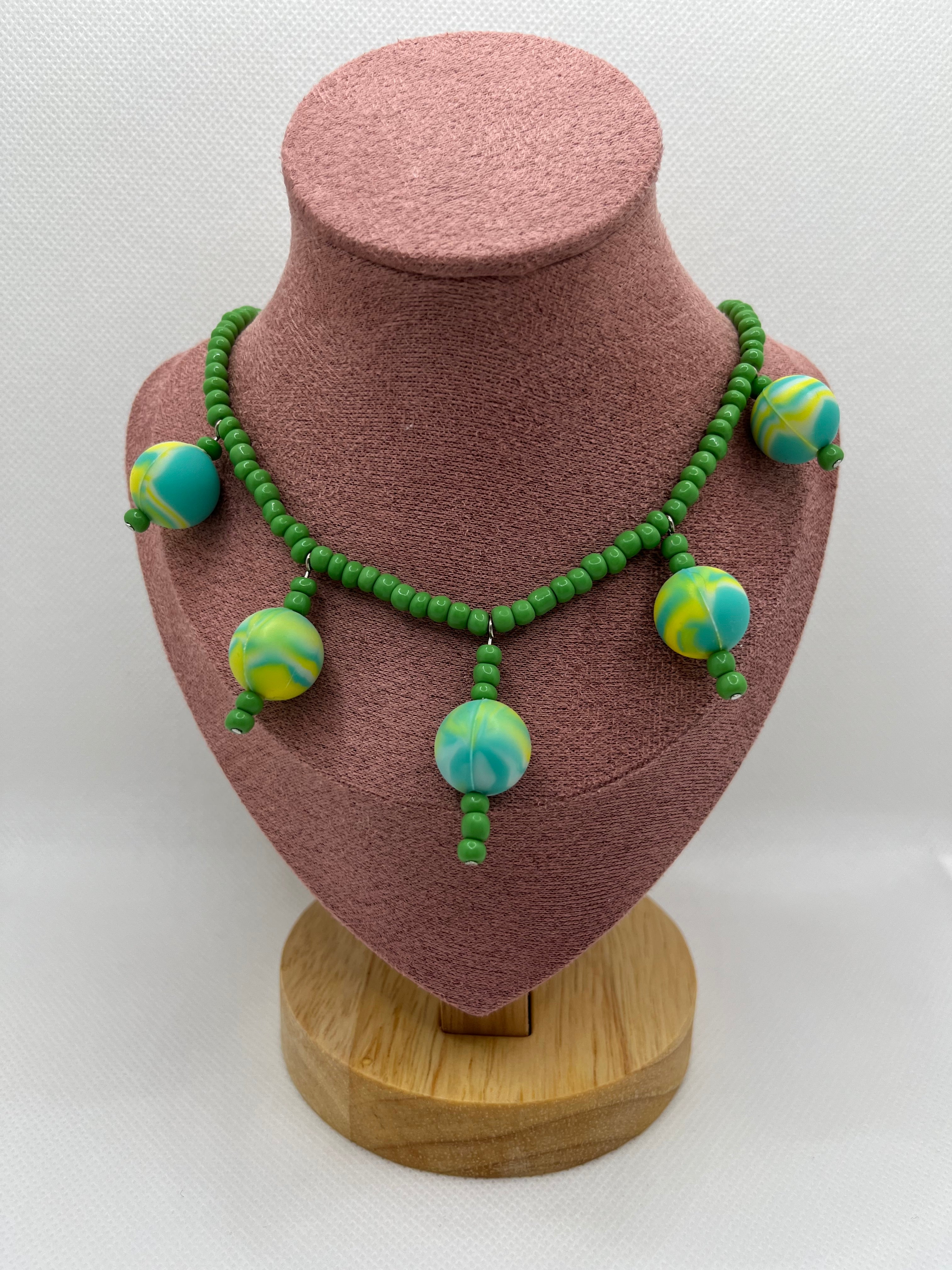 Green seed bead necklace with silicone beads strung throughout draped over a blush pink mannequin with a white background.