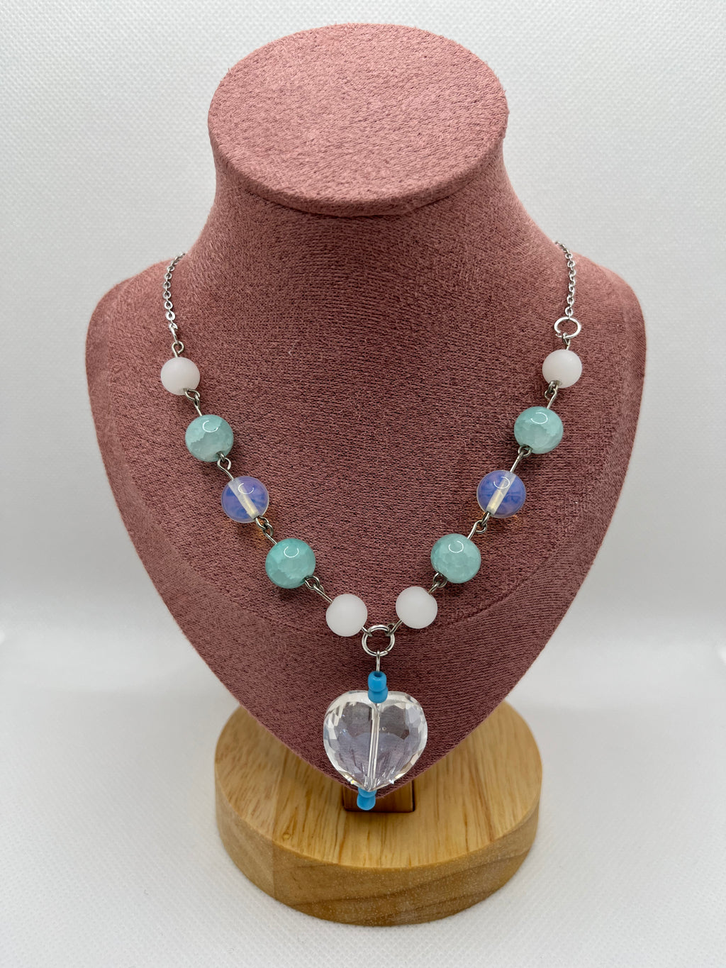 Blue, white, and clear necklace with a heart-shaped pendant draped over a blush pink mannequin with a white background.