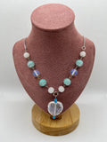 Blue, white, and clear necklace with a heart-shaped pendant draped over a blush pink mannequin with a white background.
