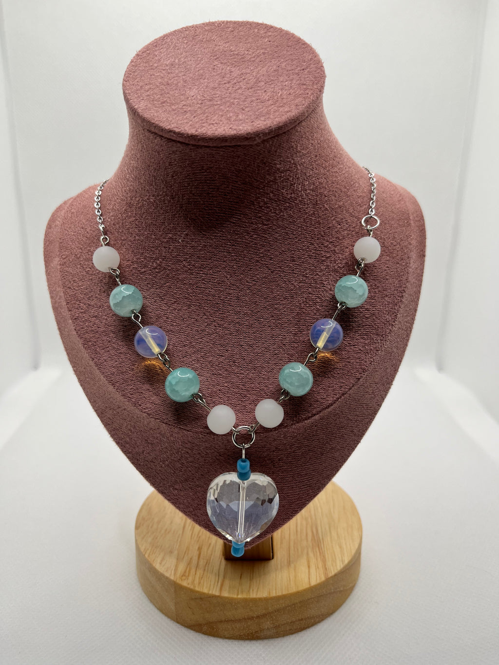 Blue, white, and clear necklace with a heart-shaped pendant draped over a blush pink mannequin with a white background.