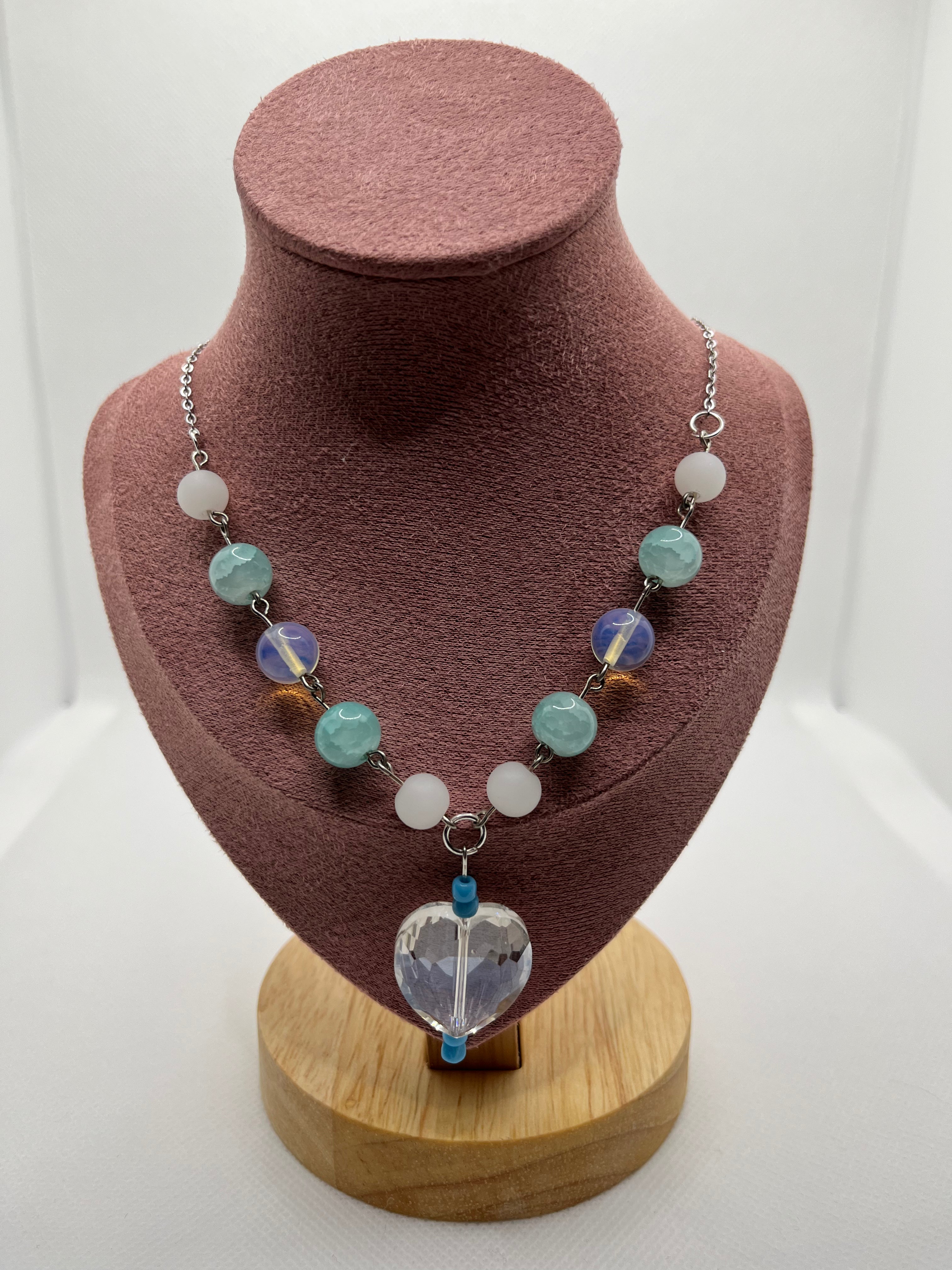 Blue, white, and clear necklace with a heart-shaped pendant draped over a blush pink mannequin with a white background.